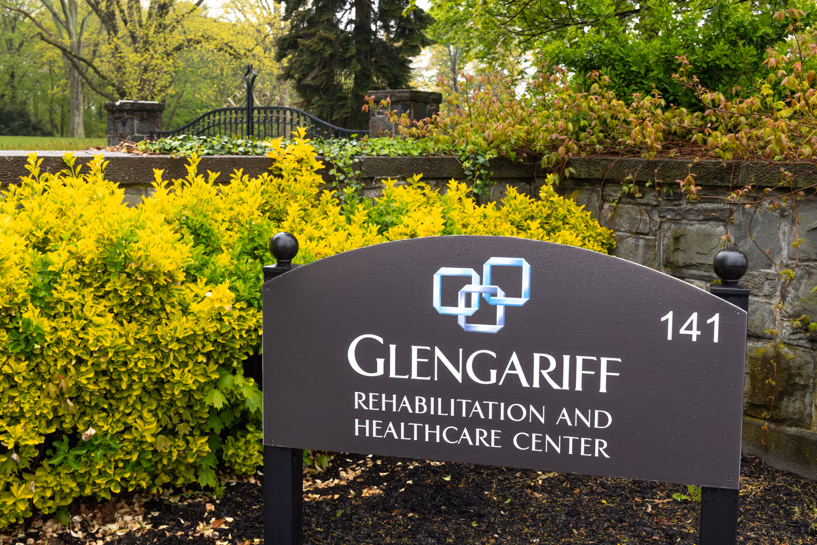 Outdoor sign for Glengariff Rehabilitation and Healthcare Center with the number 141, surrounded by green and yellow bushes and a stone wall with a black iron gate in the background.