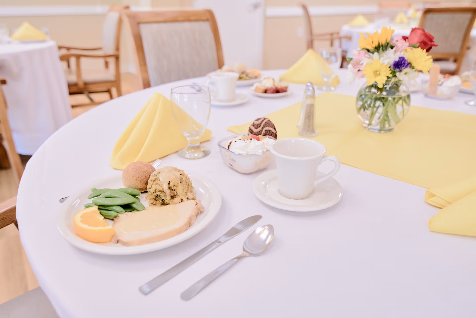 A round dining table set with a plate of food, a coffee cup, folded yellow napkins, and a vase of flowers in a dining room.