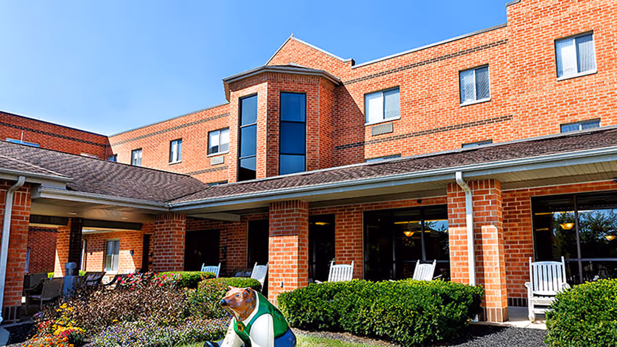 Front exterior of a red-brick senior living building with a covered porch, rocking chairs, shrubs, and a decorative animal statue.