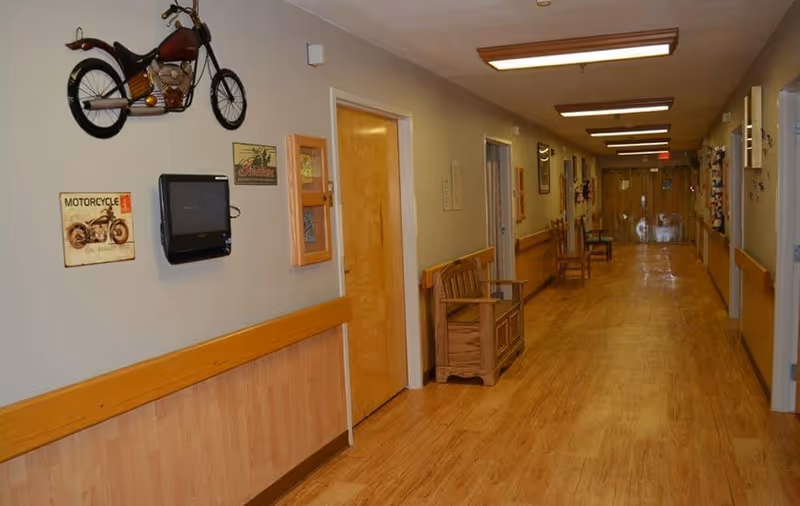 A long indoor hallway with wooden flooring and beige walls, decorated with motorcycle-themed wall art and a small mounted TV. There are wooden benches along the walls and several doors leading to rooms. The hallway is well-lit with ceiling lights and has handrails on both sides.