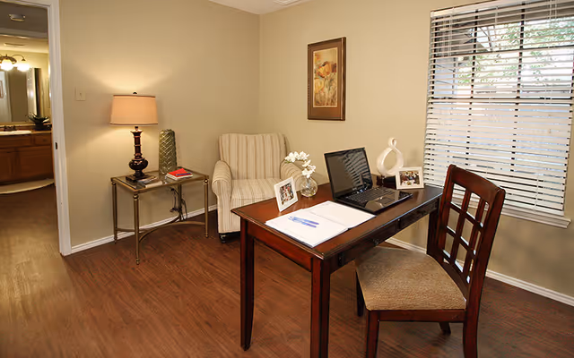 A cozy interior room with a wooden desk and chair near a window with blinds. On the desk are a laptop, a notebook with a pen, a small vase with white flowers, and two framed photos. To the left is a striped armchair next to a small side table with a lamp and decorative items. The room has wooden flooring and beige walls, with a framed floral picture hanging above the armchair. A doorway leads to a bathroom area with a sink and mirror visible.
