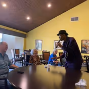 A man wearing a hat and dark clothing is performing or speaking into a microphone in front of three elderly people seated around a table in a room with a high wooden ceiling and yellow walls.