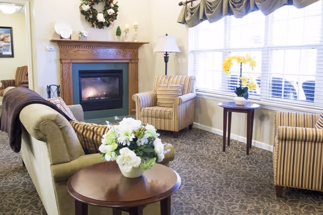 A bright sitting room with a fireplace, a sofa and striped armchairs arranged around small tables topped with floral arrangements in front of a large window.