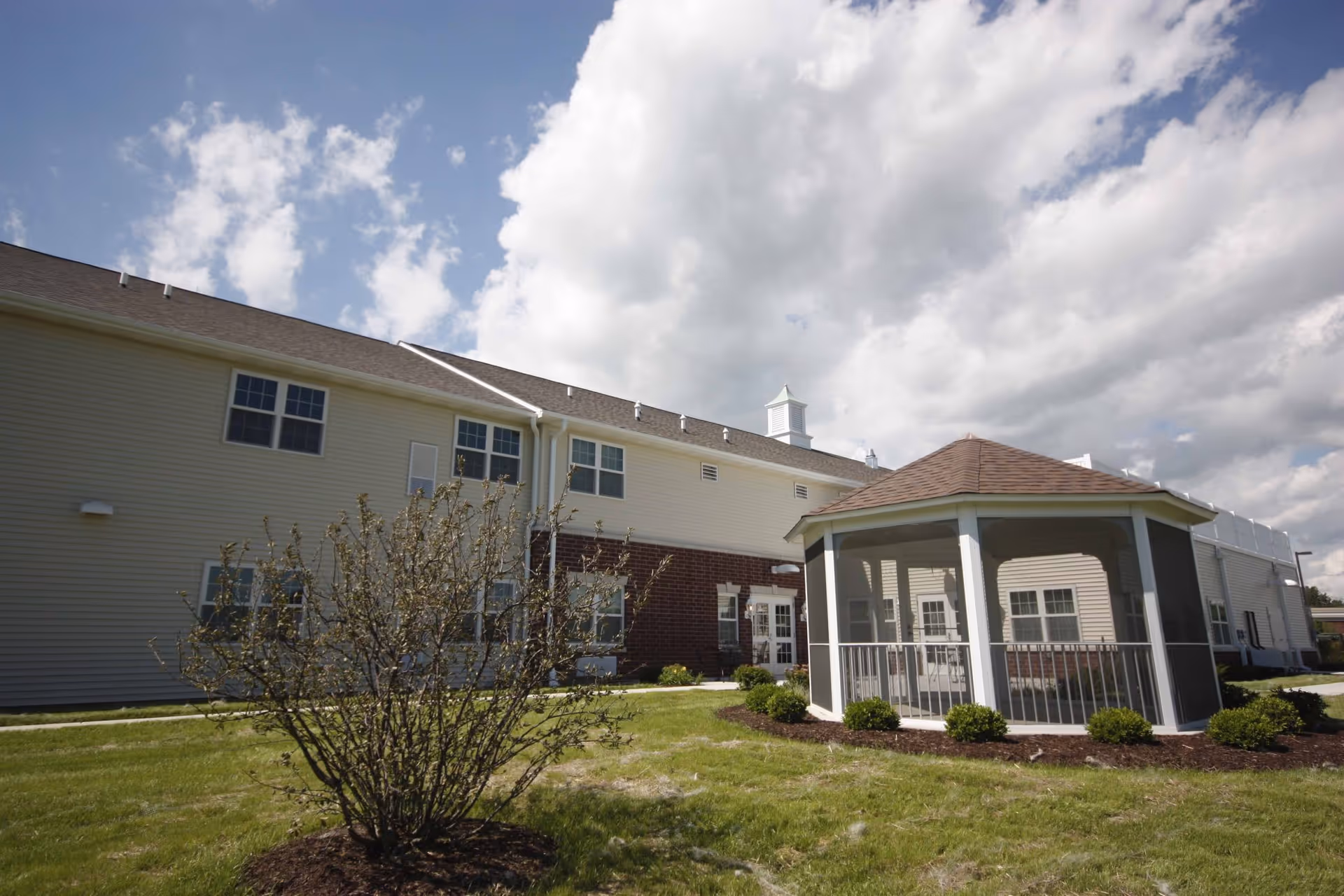 Outdoor view of a senior living facility building with beige siding and brick accents under a partly cloudy sky. In the foreground, there is a small landscaped area with a bush and a white gazebo with a brown roof surrounded by small shrubs.