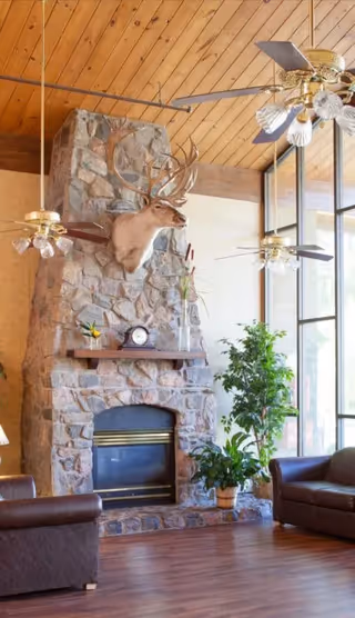 A cozy living room with a stone fireplace featuring a mounted deer head above the mantel. The mantel holds a clock and decorative items. The room has wooden floors, leather armchairs, large windows letting in natural light, ceiling fans with lights, and potted plants near the fireplace.
