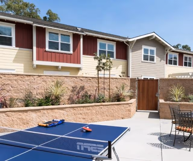 Outdoor courtyard area at Westmont of Santa Barbara featuring a blue ping pong table with paddles and balls, surrounded by a concrete patio, a wooden gate, and a two-story building with red and beige siding and multiple windows.