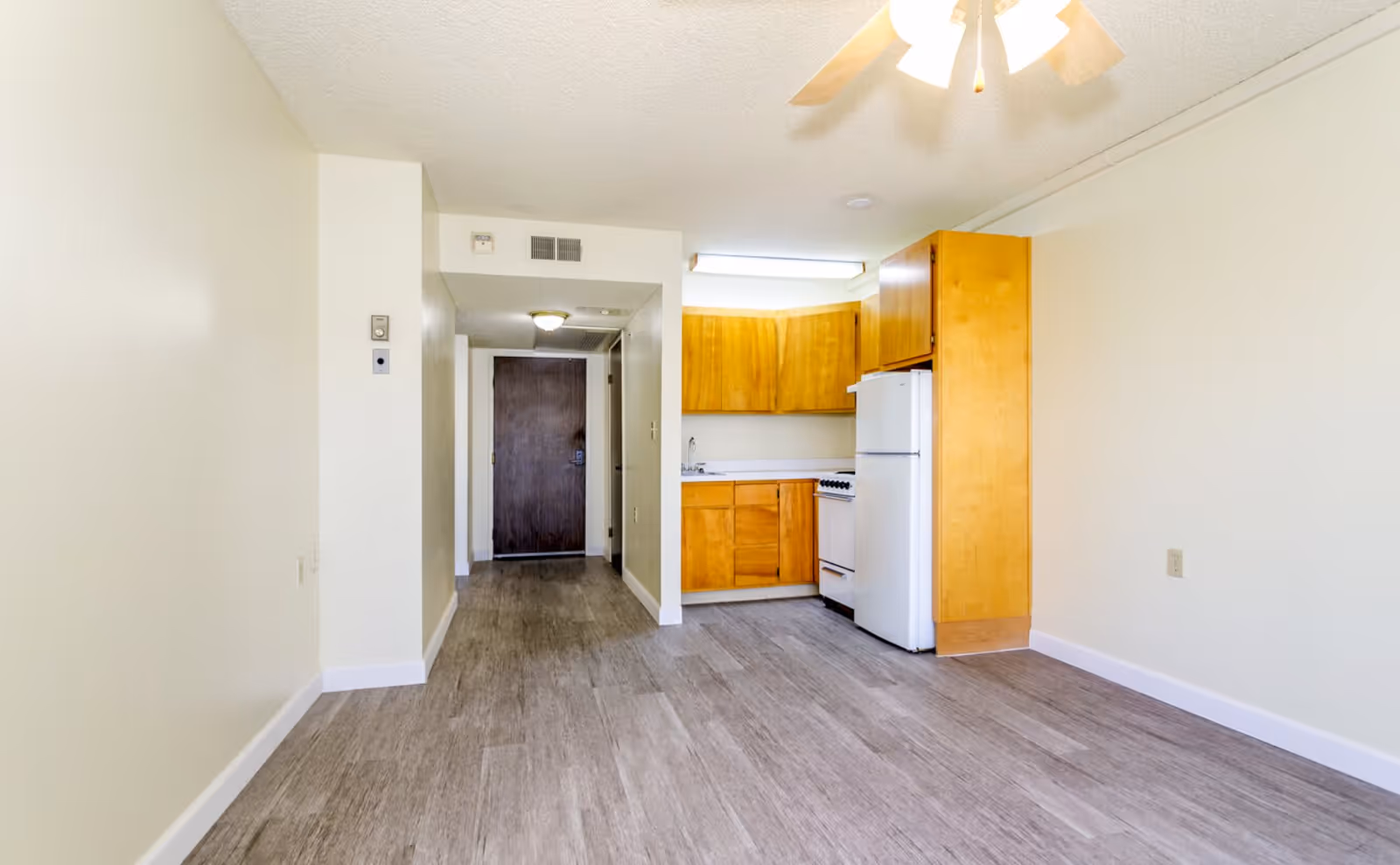 Empty interior room with light-colored walls and wood-look flooring. On the right side, there is a small kitchen area with wooden cabinets, a white refrigerator, and a stove. A ceiling fan with lights is visible above. At the far end, there is a dark wooden door and a hallway leading to it.