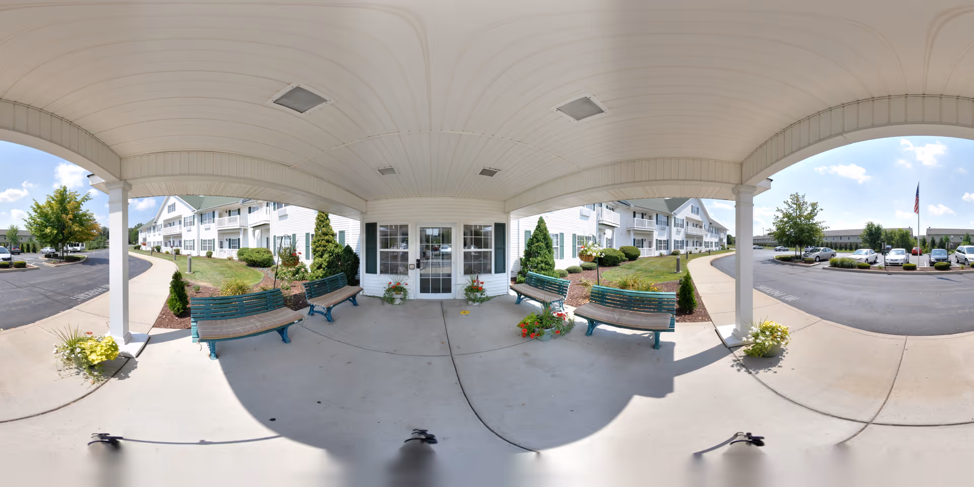 Covered entrance/portico of a white senior living building with benches, potted plants, and a drive-up area.