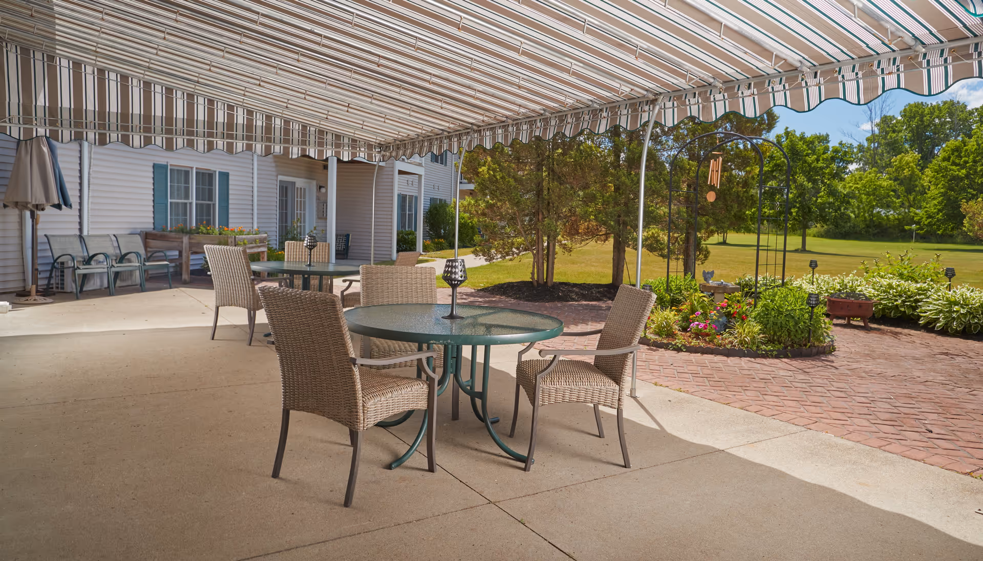 Outdoor patio area at Ashton Place Senior Living with a round glass table and four wicker chairs under a striped retractable awning. The patio overlooks a garden with flowers, shrubs, and trees, with a brick walkway and a grassy lawn in the background.