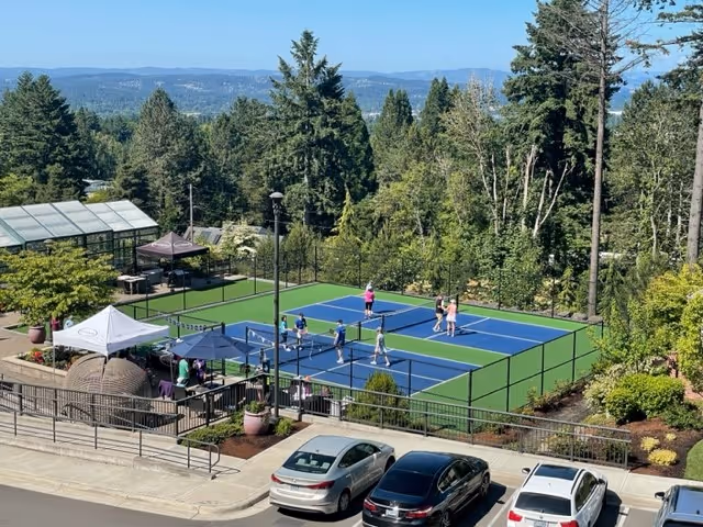 People playing on blue-and-green outdoor pickleball courts next to a parking area with cars and forested hills beyond.