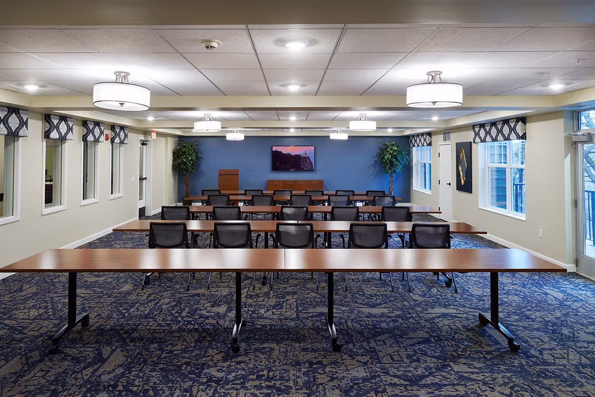 Spacious multipurpose meeting room with rows of tables and chairs facing a TV and podium against a blue accent wall.
