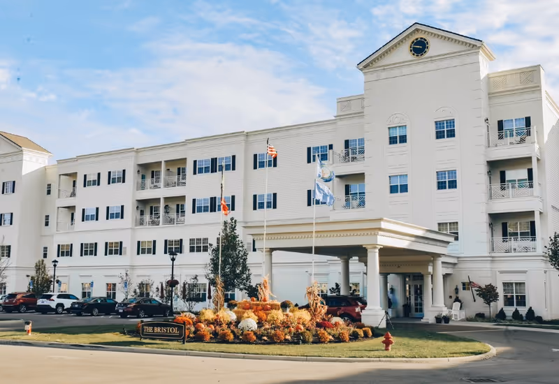 White multi-story senior living building named The Bristol at Worthington Hills with covered entrance and flags.