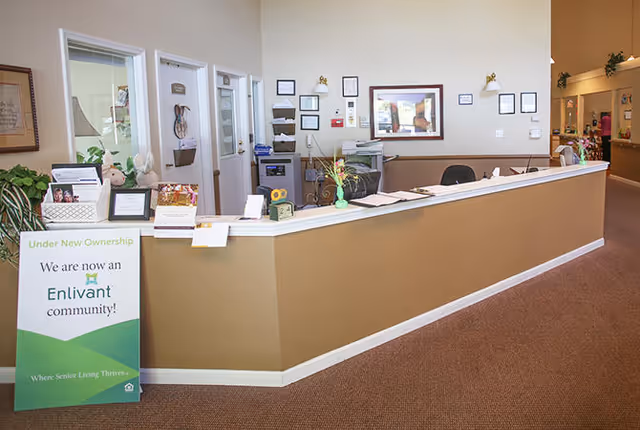 Reception desk area inside a senior living facility with a sign indicating new ownership by Enlivant. The desk has office supplies, a computer, and decorative plants. The background shows doors, framed certificates, and a hallway leading to other rooms.