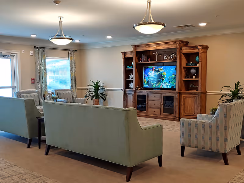 A senior living facility common area with light green sofas and patterned armchairs arranged around a large wooden entertainment center with a flat-screen TV displaying an underwater scene. The room has beige walls, carpeted floor, two ceiling light fixtures, potted plants, and a window with patterned curtains.