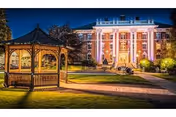 Illuminated historic brick building front at dusk with a lit wooden gazebo and paved walkways on a manicured lawn.