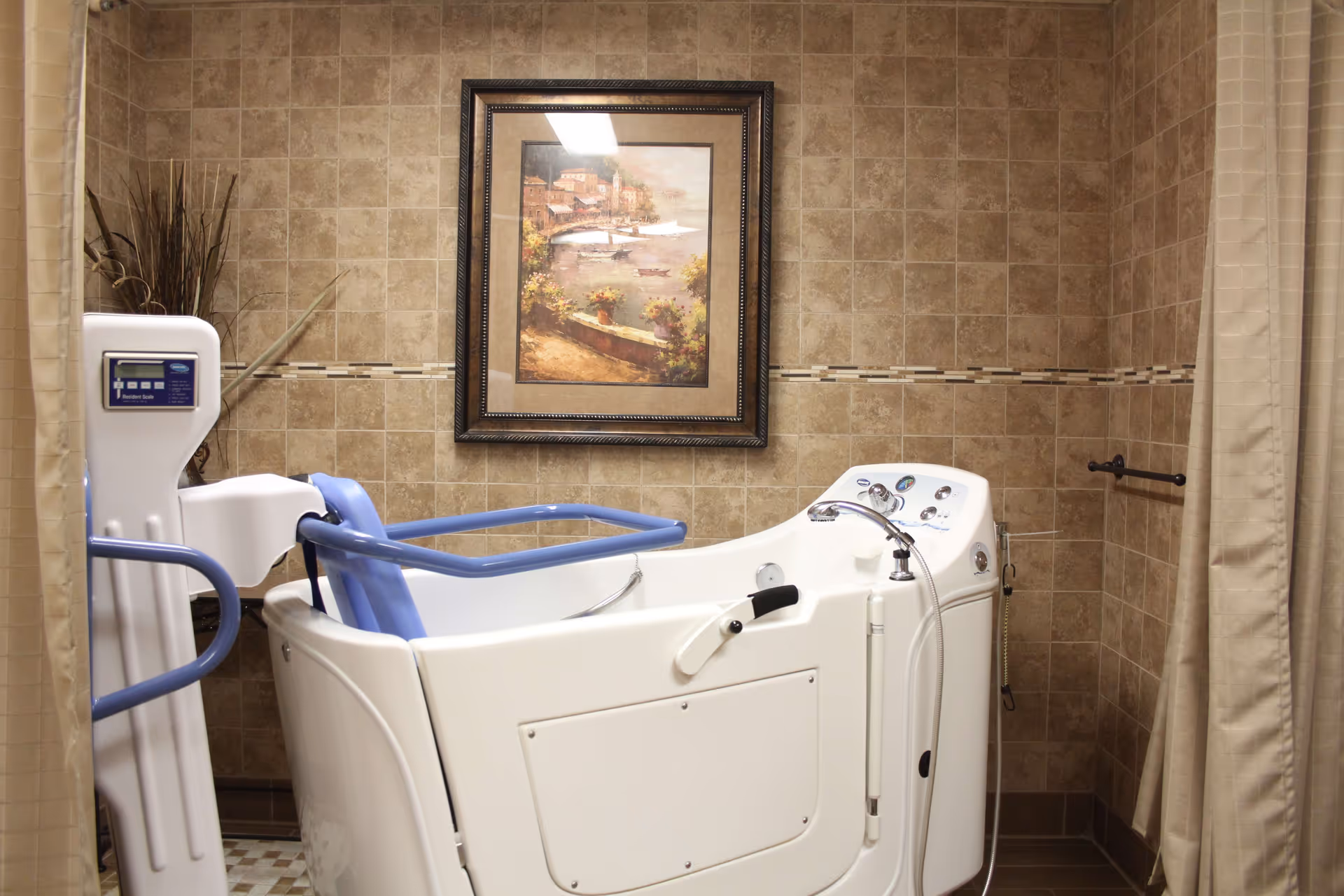 A bathroom with beige tiled walls featuring a walk-in bathtub with blue handrails and a control panel. There is a framed painting of a coastal scene hanging on the wall above the bathtub, and a beige shower curtain is partially visible on the right side.