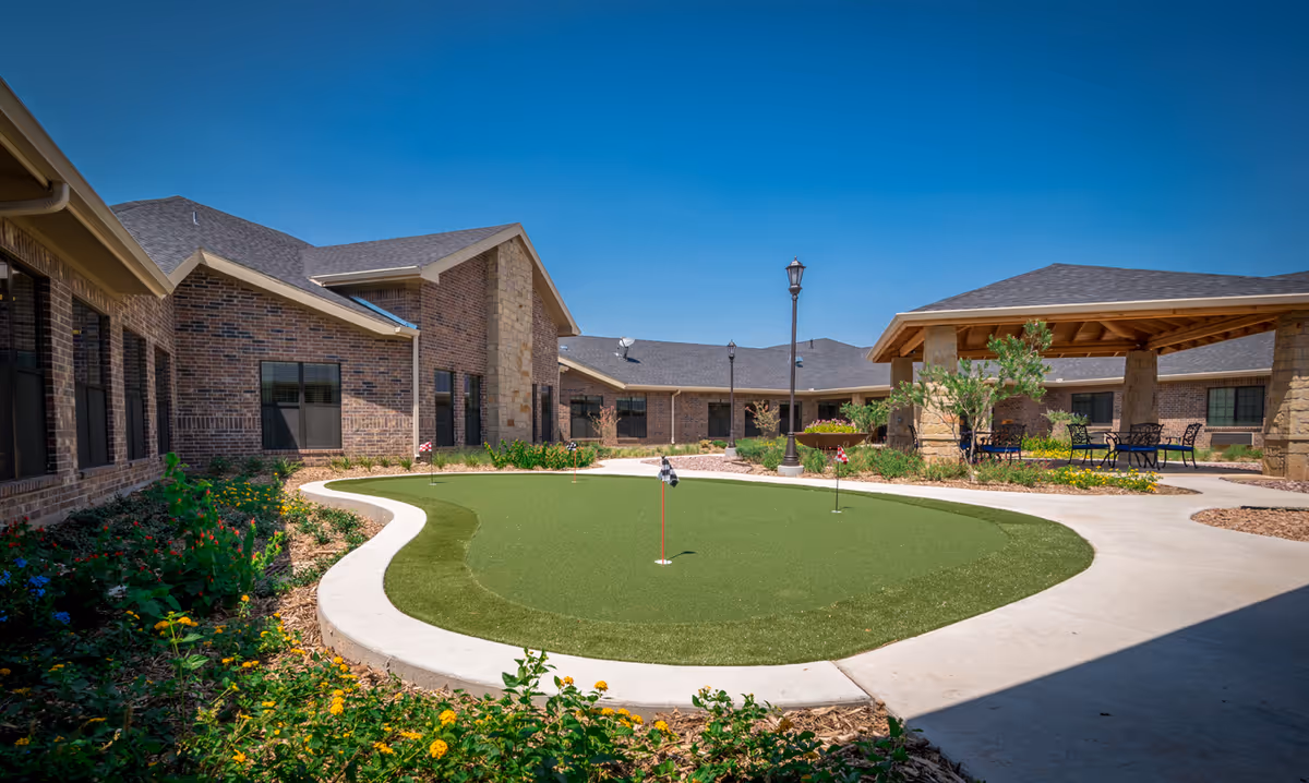 Outdoor courtyard area at Ashton Medical Lodge featuring a putting green with small flags, surrounded by a curved concrete walkway, flower beds, and brick buildings under a clear blue sky. There is also a covered seating area with tables and chairs.