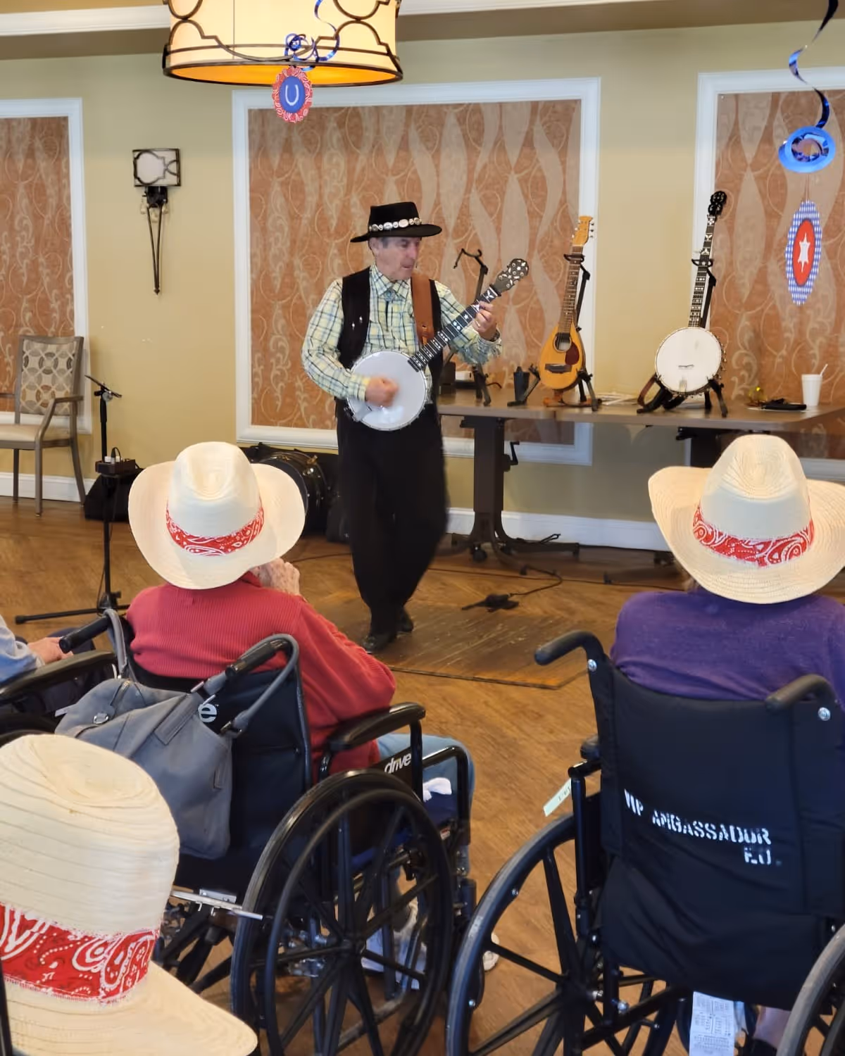 A man wearing a black hat and vest plays a banjo in front of an audience of elderly people in wheelchairs, some wearing white hats with red bandanas. The setting appears to be a decorated indoor room with musical instruments on a table behind the performer.