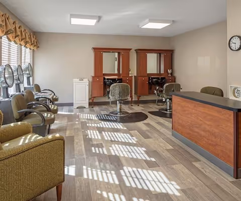A bright salon area with two wooden styling stations featuring mirrors and chairs, a row of hair drying chairs with hooded dryers along the left wall, a reception desk on the right, and large windows with blinds casting shadows on the wooden floor.