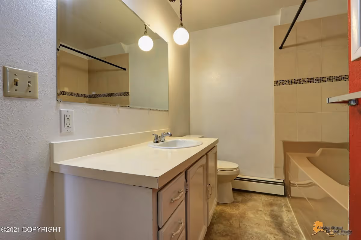 A bathroom with a white countertop and sink, a large mirror above the sink, a toilet, and a bathtub with beige tiles and a decorative tile border. The floor has a patterned linoleum surface, and there are two round pendant lights hanging from the ceiling.