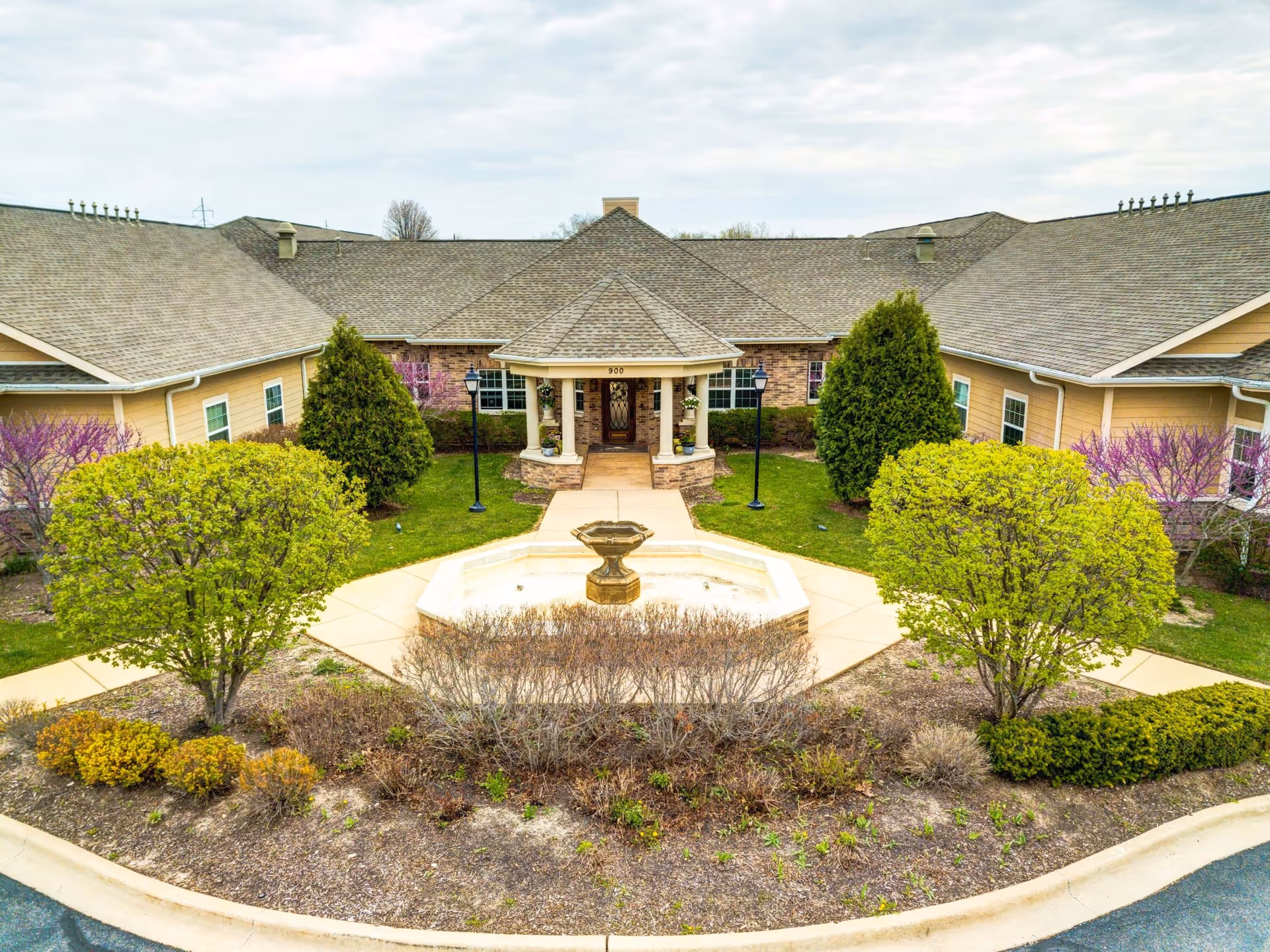 Entrance courtyard with a central fountain, landscaped greenery, and a covered portico of a single-story senior living building.