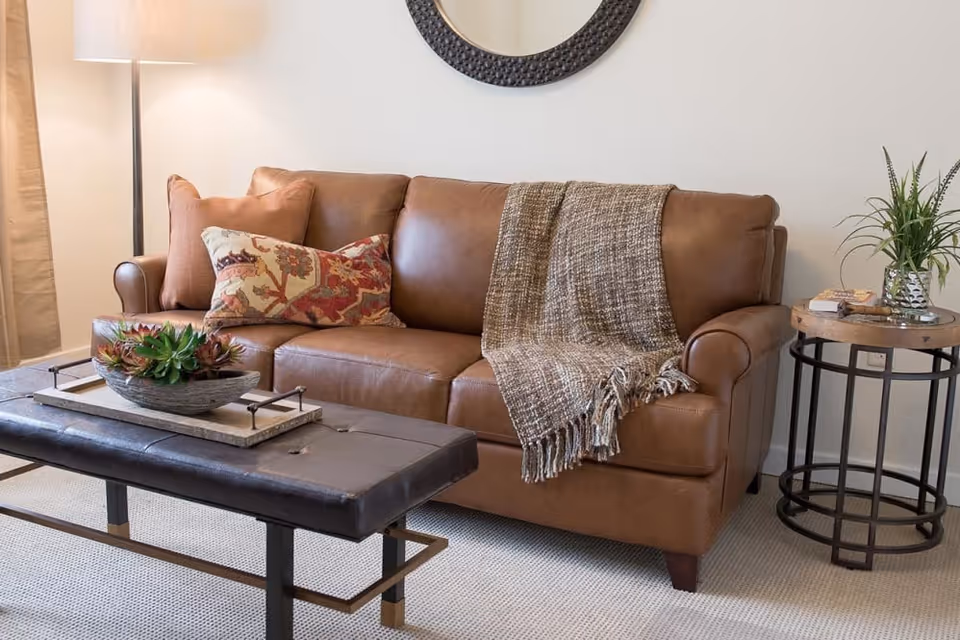 A cozy living room area featuring a brown leather sofa adorned with a patterned pillow and a textured throw blanket. In front of the sofa is a black leather-topped coffee table with a decorative tray holding a succulent plant. To the right of the sofa is a round wooden side table with a potted plant and some books. A floor lamp with a white shade stands to the left, and a round mirror hangs on the wall above the sofa.