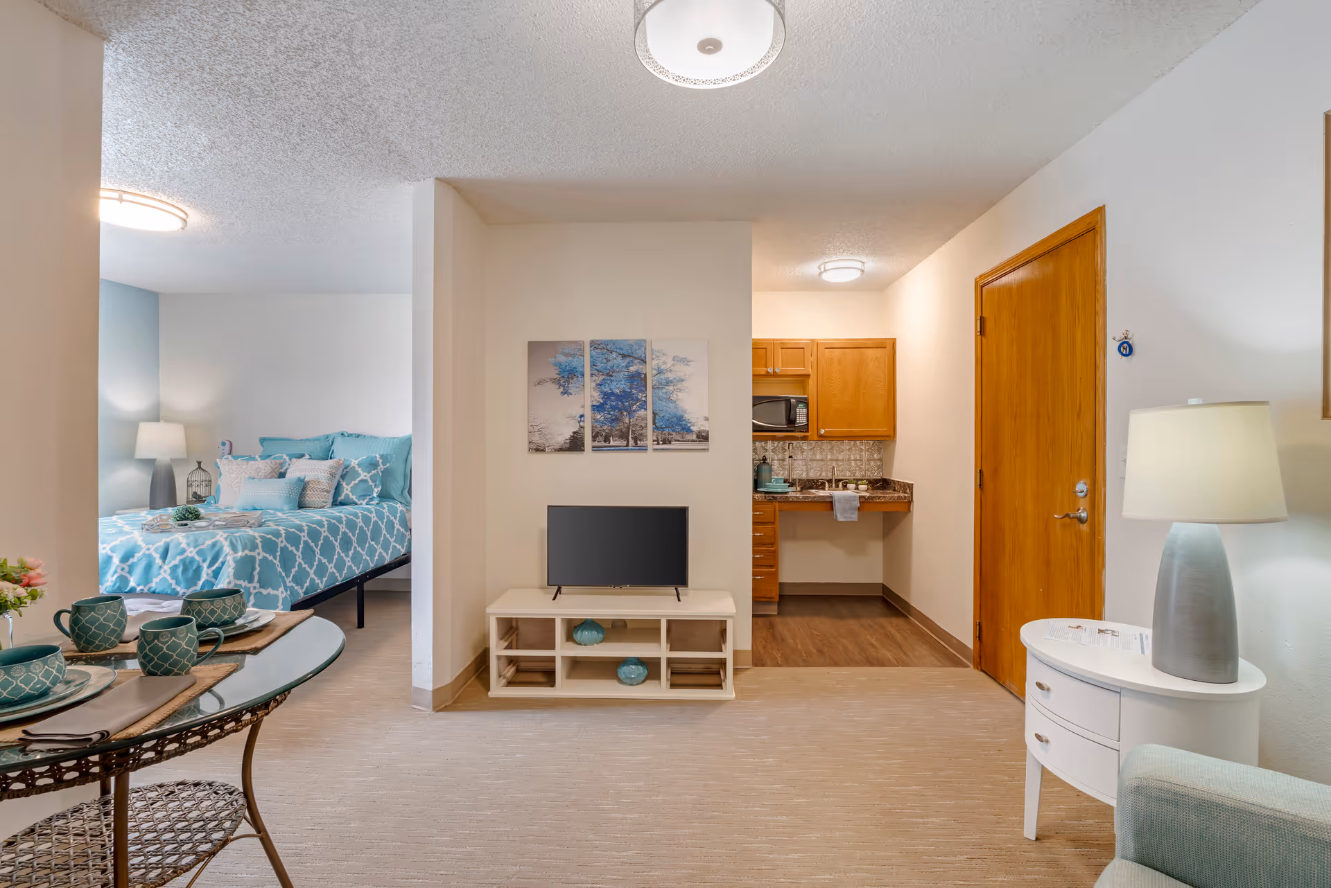 Interior view of a senior living apartment showing a small living area with a TV on a white stand, a kitchenette with wooden cabinets and a microwave, and a bedroom with a bed dressed in blue and white patterned bedding. A round glass dining table with teal dishware is partially visible on the left, and a white side table with a lamp is on the right.