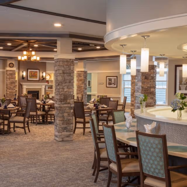 A spacious dining area in a senior living facility featuring multiple round tables with chairs arranged around them. The room has stone pillars, soft lighting with hanging pendant lights, and a cozy fireplace in the background. The decor includes framed artwork on the walls and floral centerpieces on the tables.
