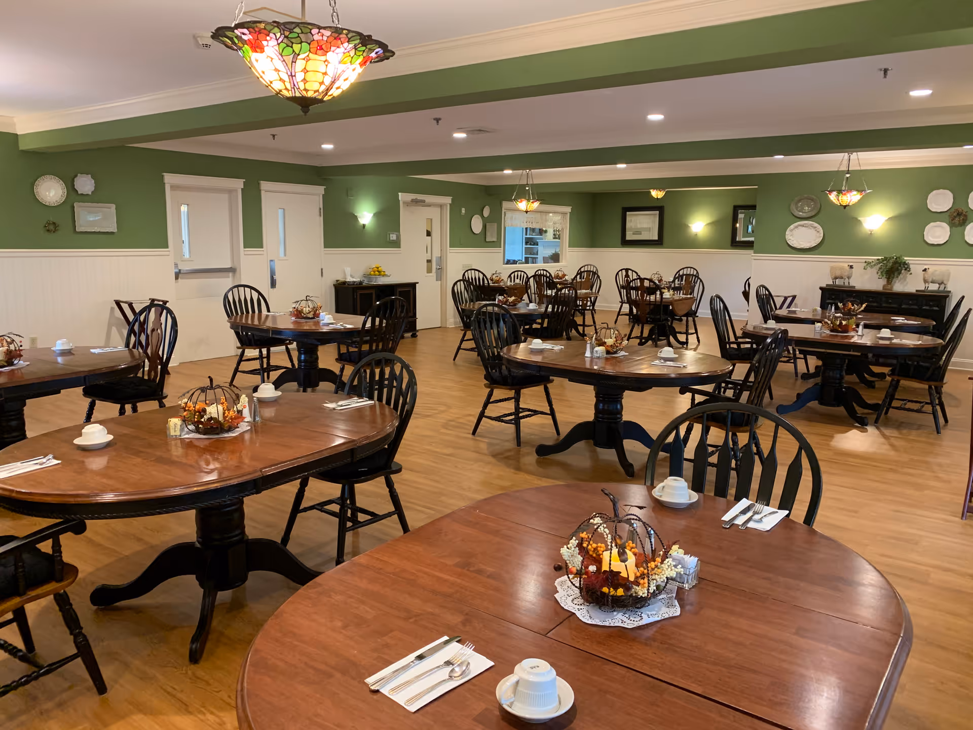 A bright dining room with multiple wooden tables and chairs, green walls, decorative hanging lights and autumn centerpieces on each table.