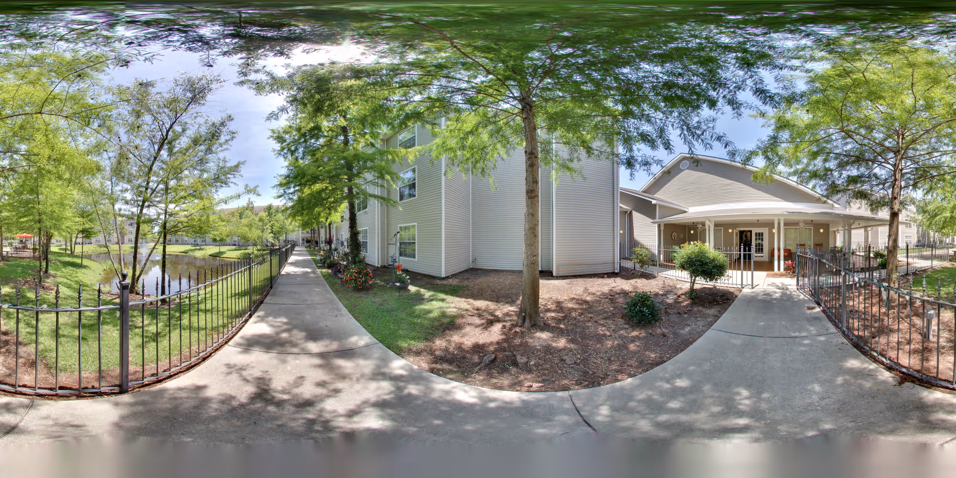 Paved walkway beside a fenced pond leading to the entrance of a senior living building surrounded by trees and landscaping.