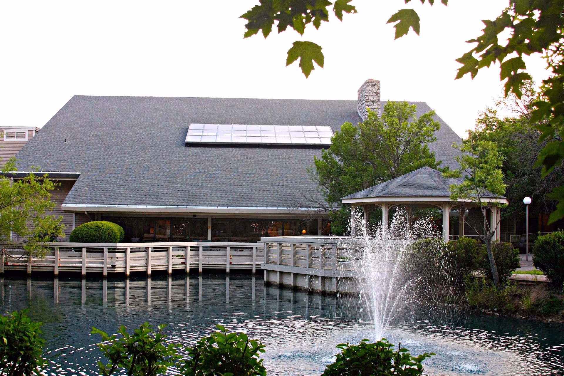 A large sloped-roof building with a wooden boardwalk, gazebo and a fountain spraying water in a pond in front.