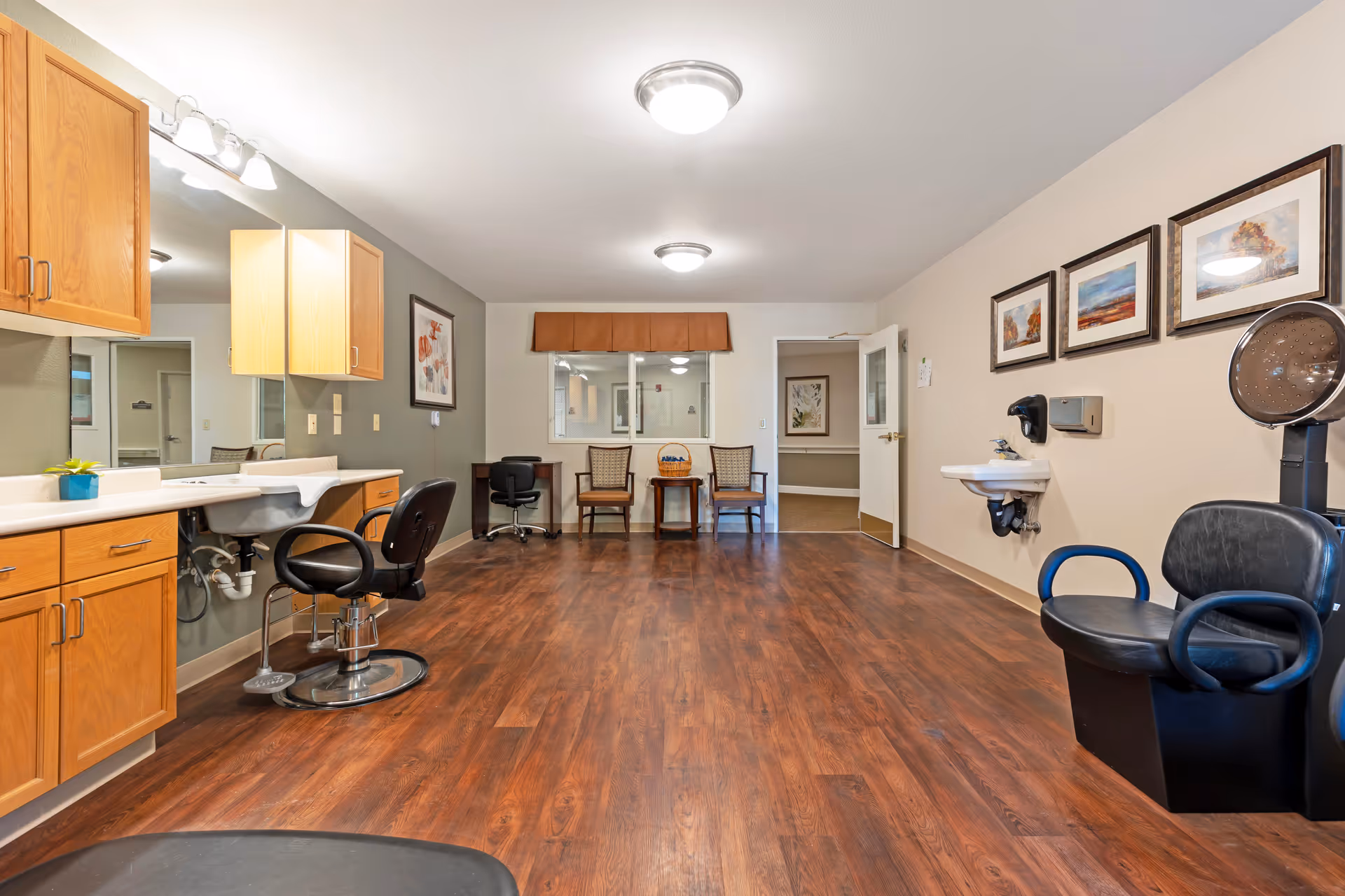 Interior view of a salon area in a senior living facility with wooden flooring, salon chairs, a hair washing station, a sink mounted on the wall, wooden cabinets, framed artwork on the walls, and a small seating area with two chairs and a table near a window.
