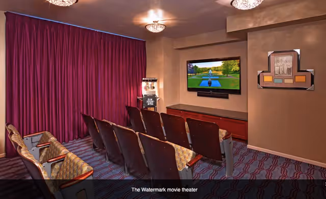 A small movie theater room with two rows of theater-style seats facing a wall-mounted flat screen TV. The room has patterned carpet, beige walls, and purple curtains covering one wall. There is a popcorn machine in the corner and a framed picture on the wall.