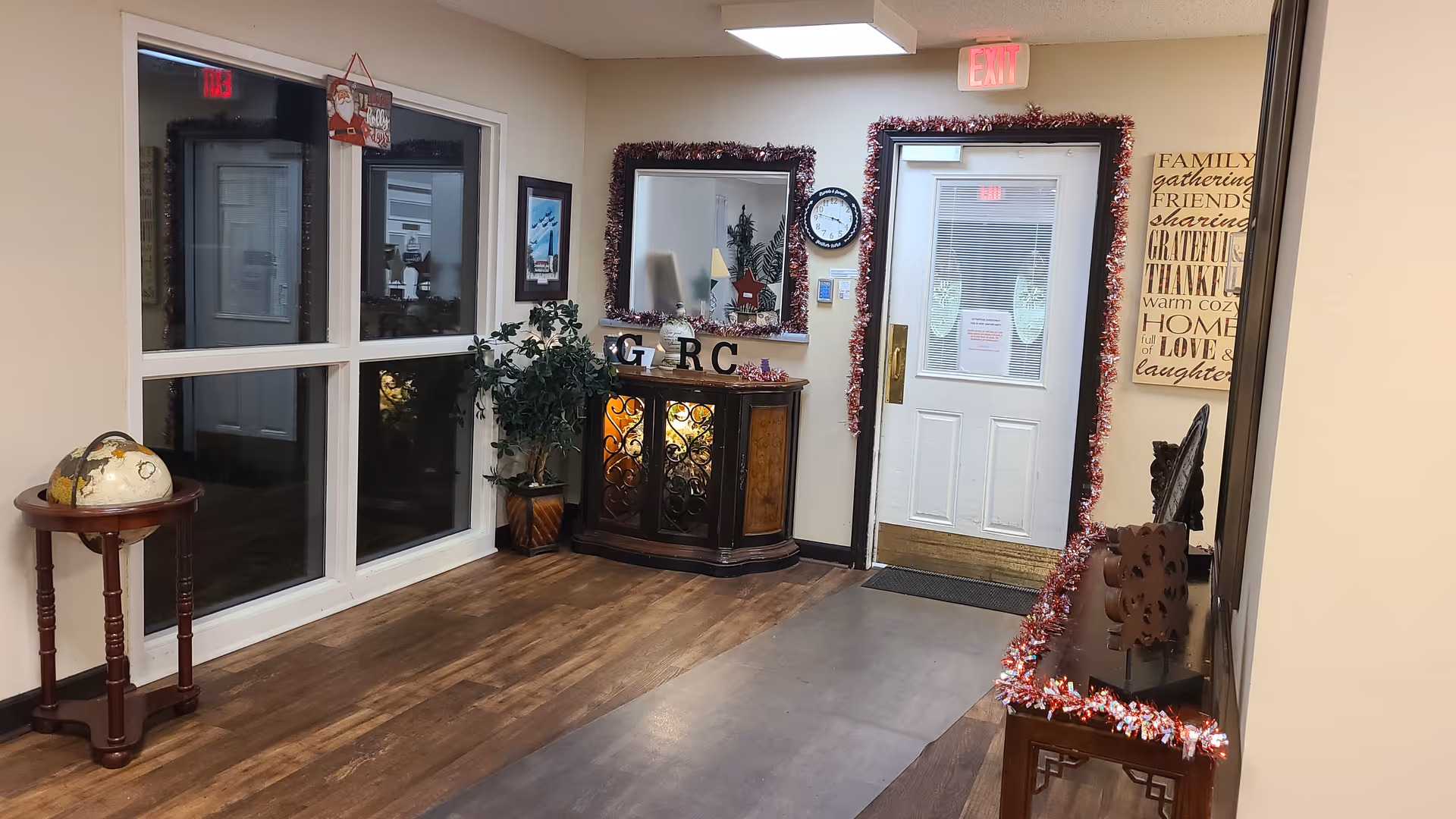 Interior hallway of a retirement center decorated with holiday garlands around a door and mirror. There is a small wooden table with a globe on the left, a cabinet with decorative items and letters 'GRC' in the center, and a wall clock above it. A framed inspirational sign is on the right wall.
