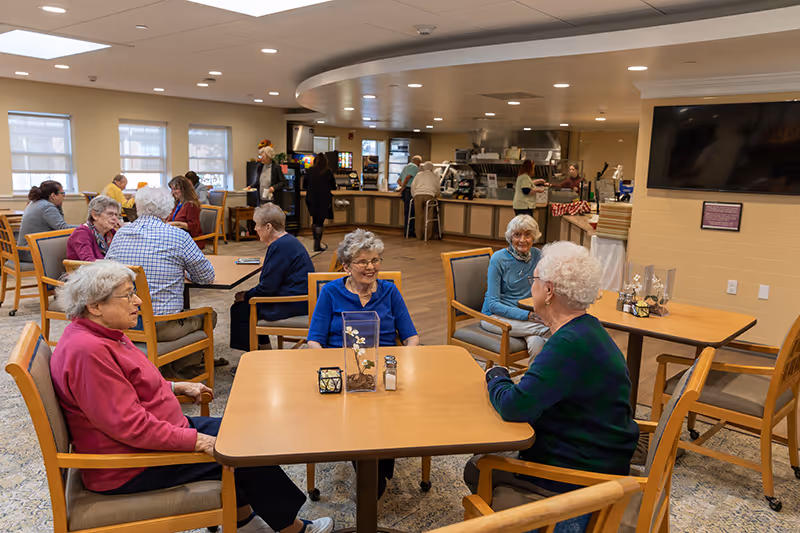 A group of elderly residents sit and chat at tables in a bright communal dining area with a serving counter and staff in the background.