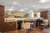Interior view of a salon area in a senior living facility with multiple salon chairs, mirrors, and cabinets. The room has wooden flooring, a clock on the wall, framed pictures, and large windows allowing natural light.