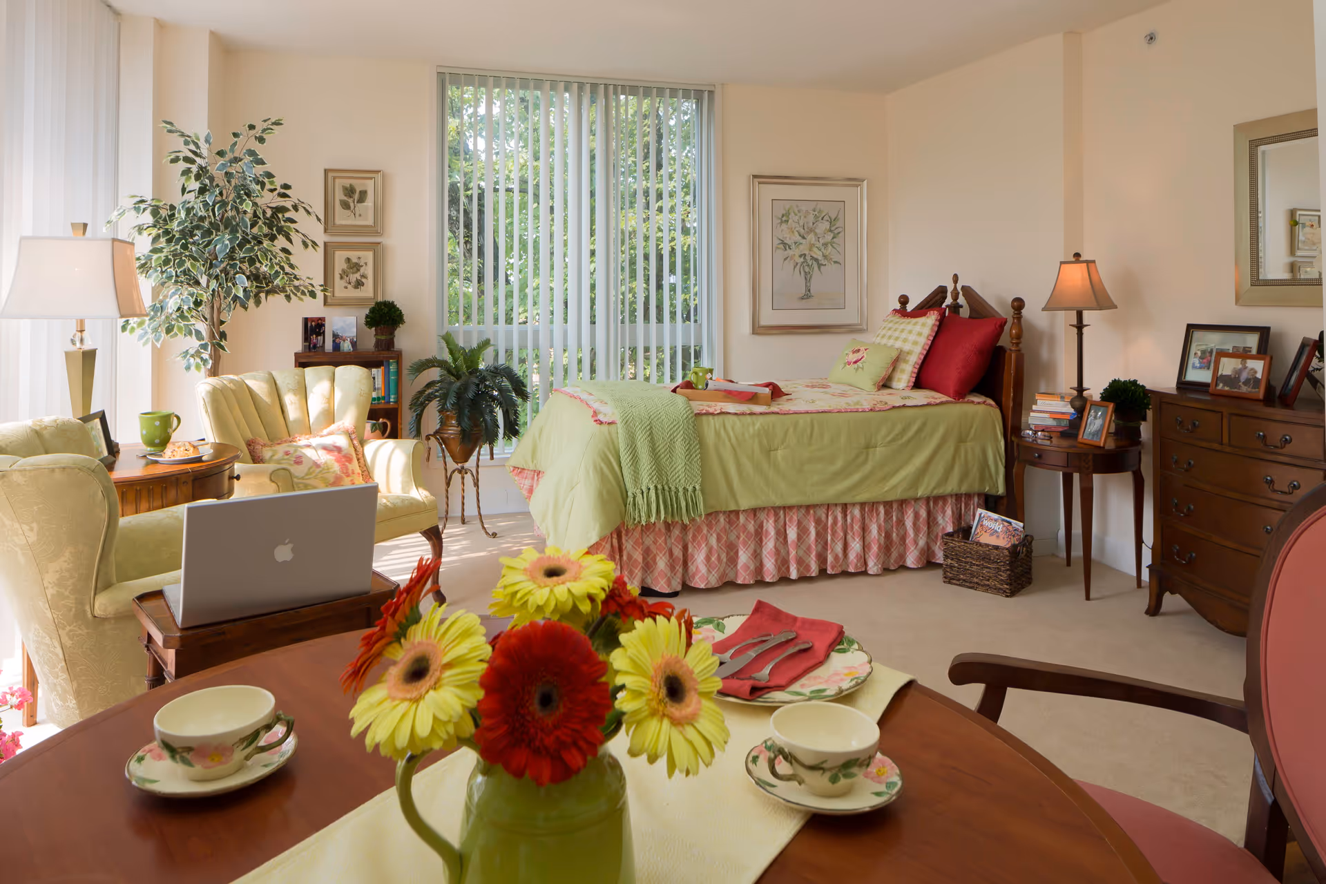 A cozy and well-lit bedroom with a single bed dressed in green and pink bedding, a wooden headboard, and several pillows. The room features a round wooden table with a floral centerpiece and tea cups, two upholstered armchairs, a laptop on a small side table, a wooden dresser with framed photos, a floor lamp, and large windows with vertical blinds letting in natural light. There are also decorative plants and framed artwork on the walls.