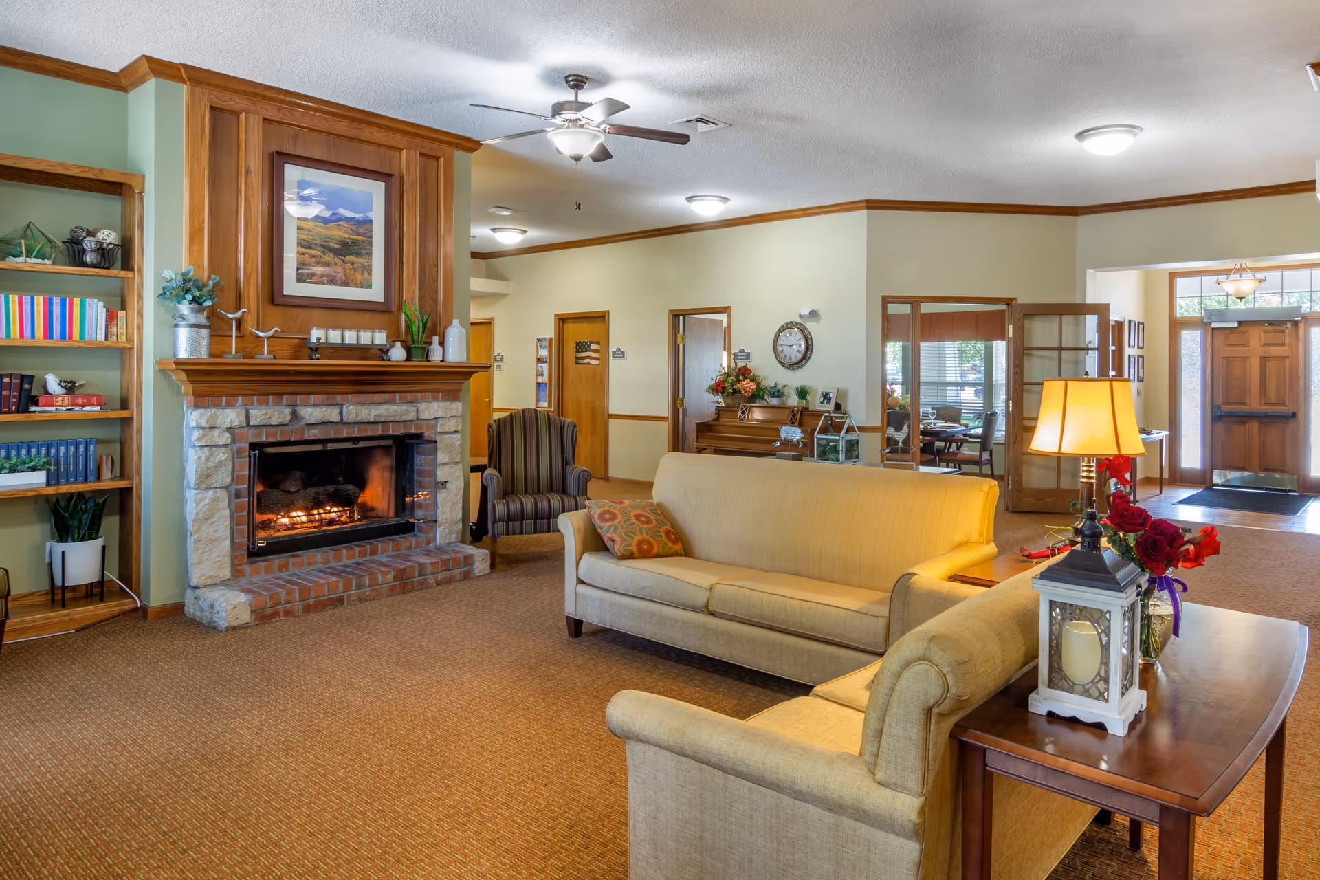 Cozy lounge with sofas, a lit stone fireplace, bookshelves, and an entryway in the background.