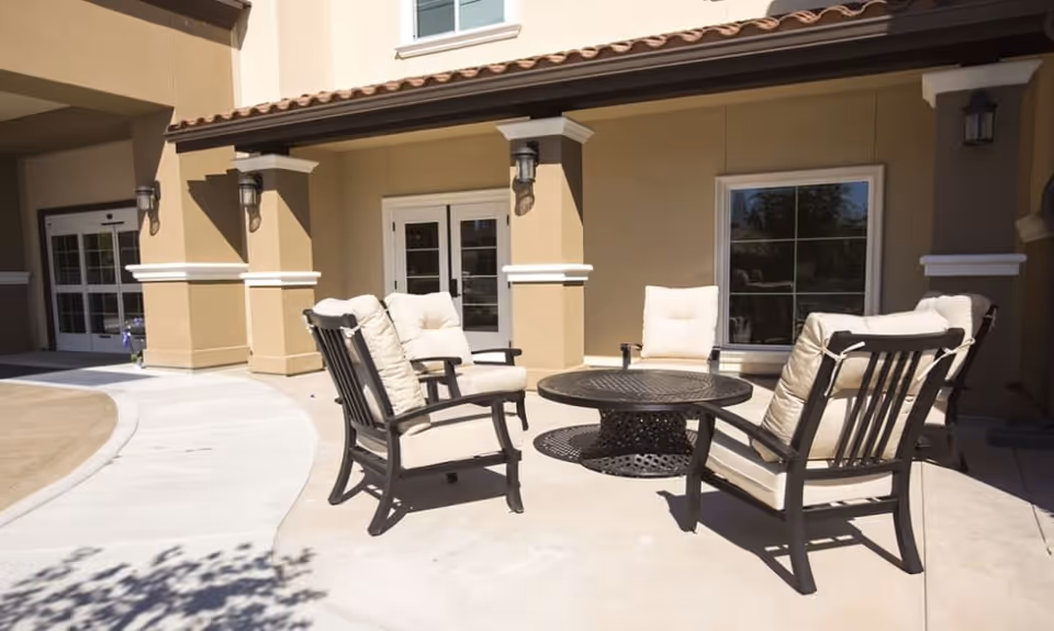 Outdoor patio area with four cushioned chairs arranged around a round metal table in front of a beige building with windows and wall-mounted lights.