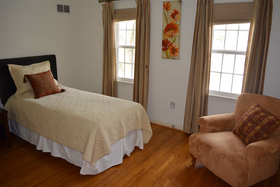 A simple bedroom with a single bed covered in a beige quilt and two pillows, one beige and one rust-colored with a pattern. There are two windows with beige curtains, a floral painting on the wall between the windows, and a brown upholstered armchair with a patterned cushion. The floor is wooden.