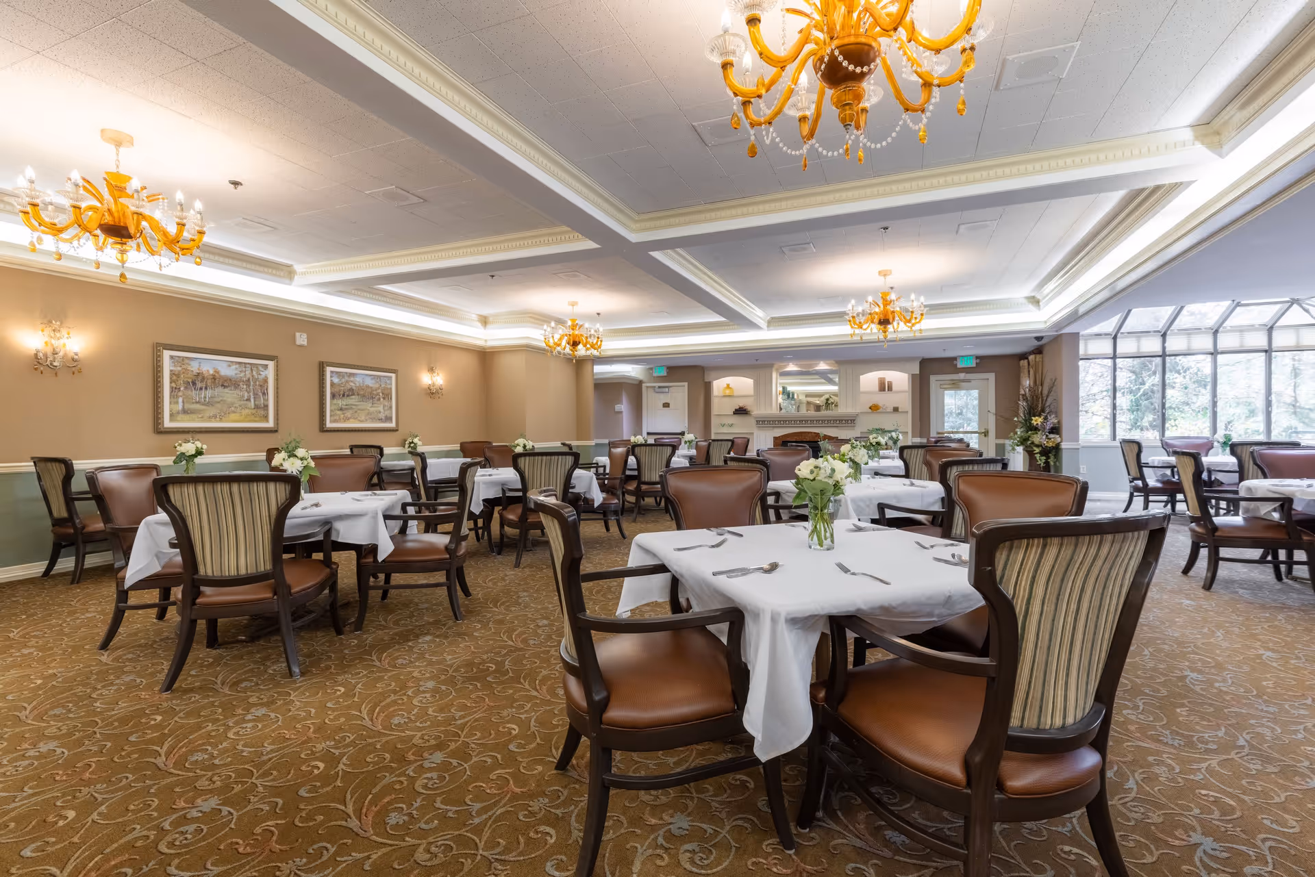 A spacious dining room with multiple tables covered in white tablecloths, each set with silverware and a vase of white flowers. The room features elegant chandeliers, framed artwork on the walls, and large windows allowing natural light to fill the space.