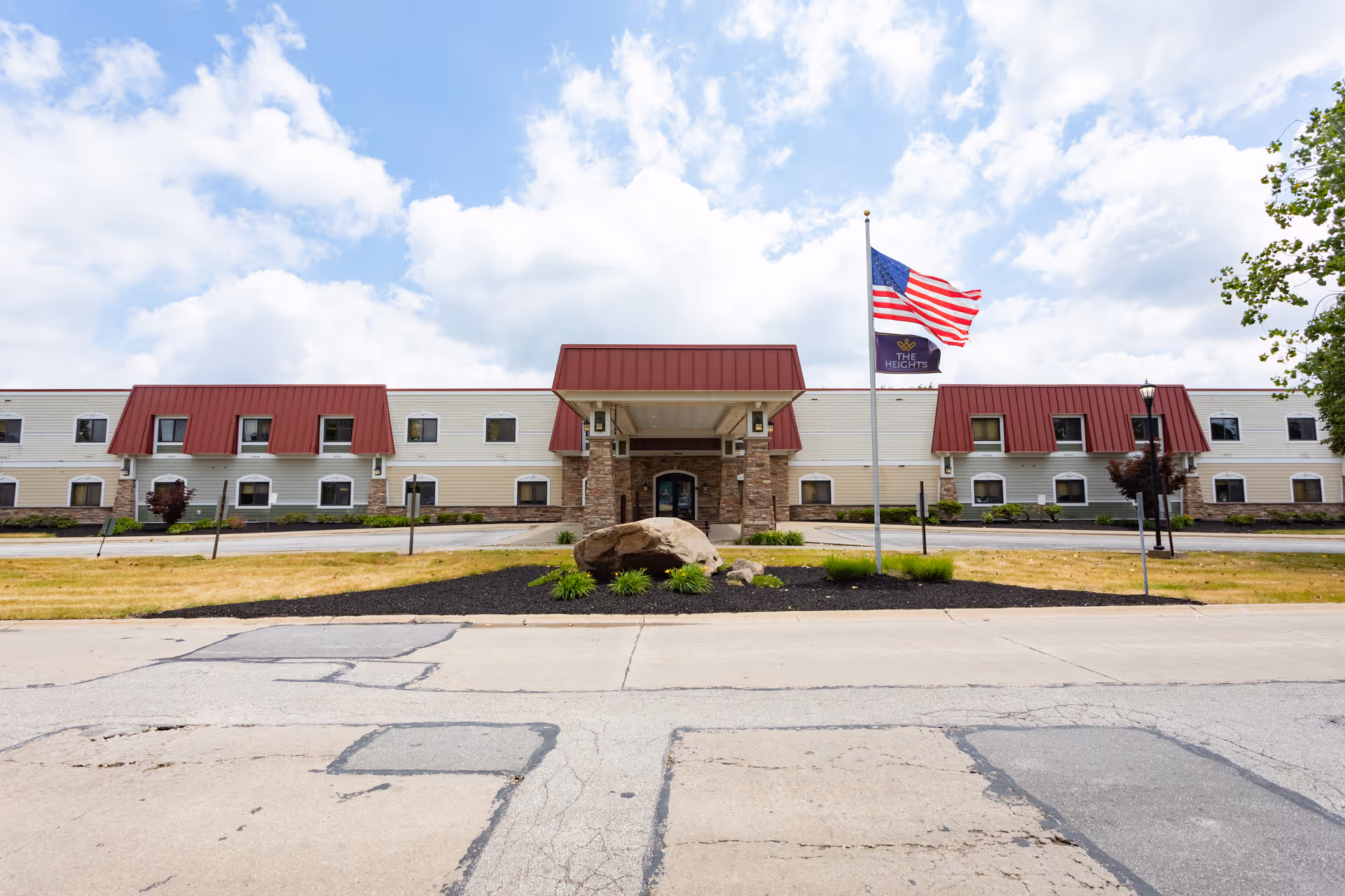 Front exterior view of The Heights Rehabilitation and Healthcare Center building with a two-story structure, red roof accents, an entrance canopy supported by stone pillars, an American flag and a facility flag flying on flagpoles, and a partly cloudy sky in the background.