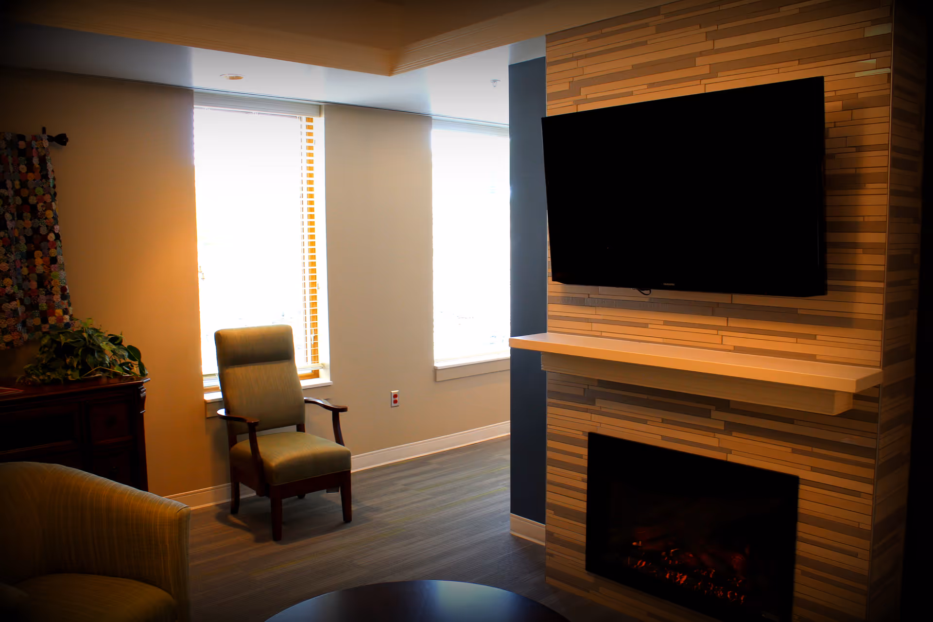 A cozy living room area with a mounted flat-screen TV above a modern electric fireplace. There are two chairs, one green armchair and one wooden armchair with green upholstery, positioned near two large windows with blinds. A wooden cabinet with a plant and a colorful quilt hanging on the wall is visible on the left side.