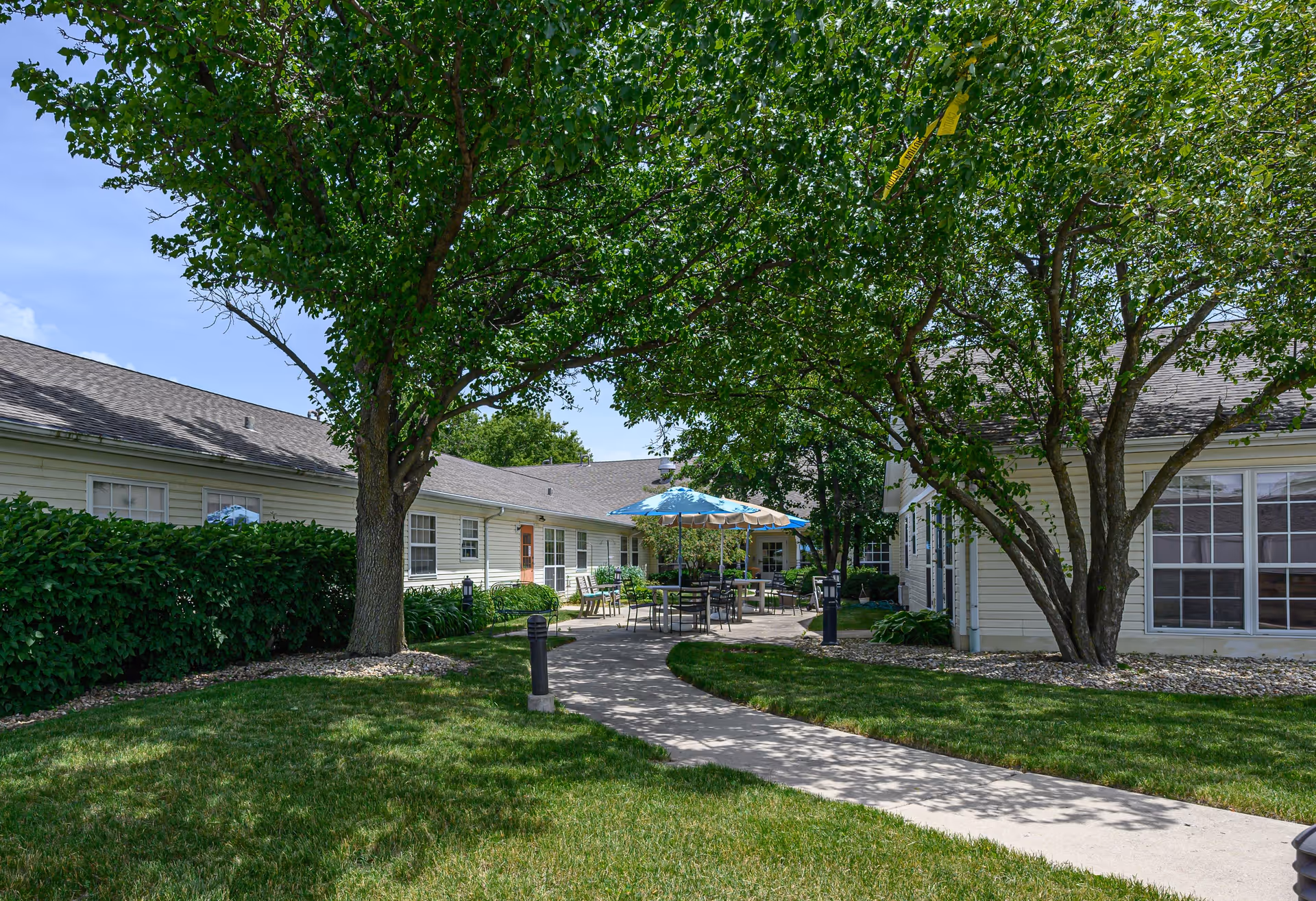 Outdoor courtyard area at Arden Courts - ProMedica Memory Care Community (Elk Grove) featuring a paved walkway, green grass, trees providing shade, and patio tables with umbrellas for seating.