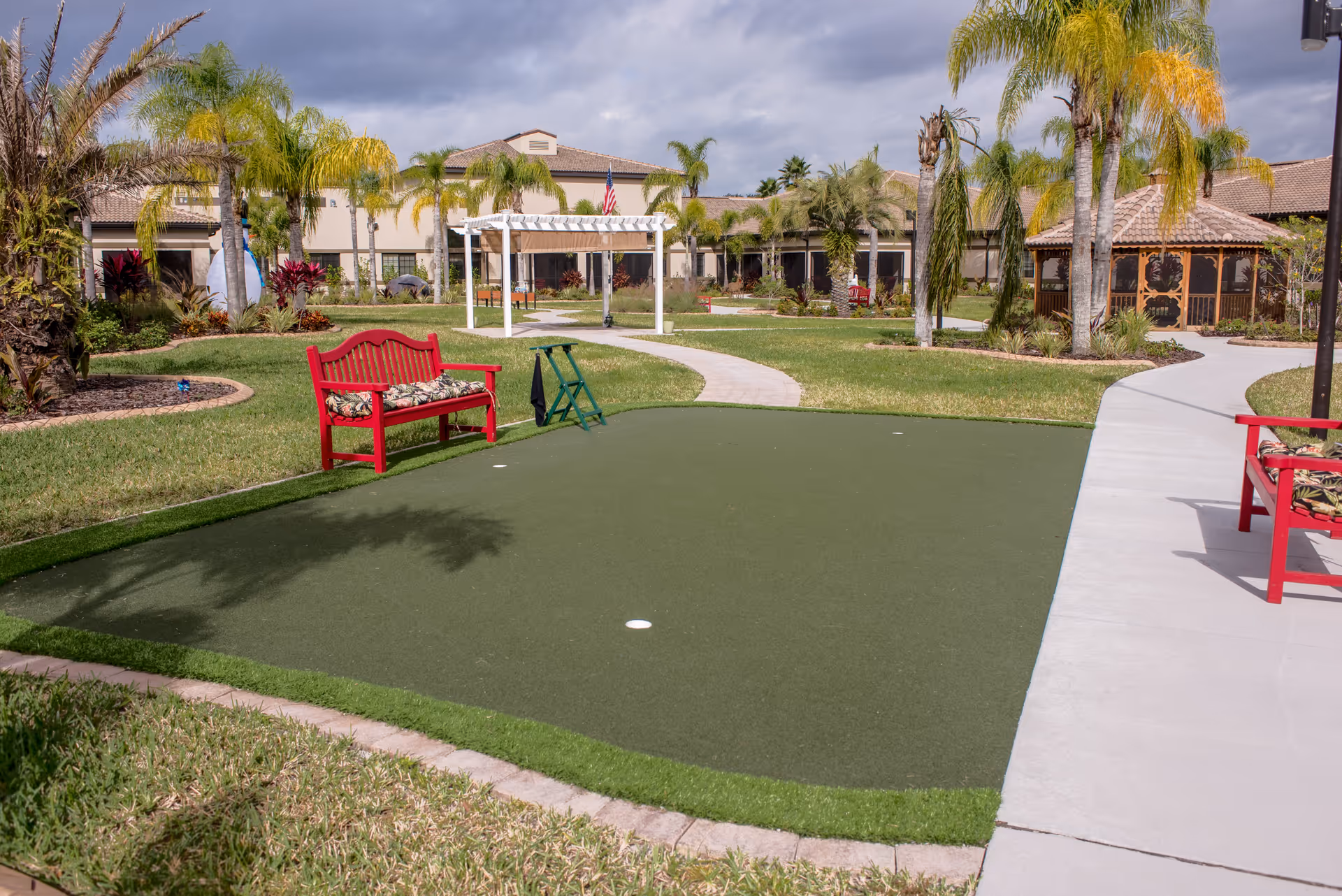 Outdoor putting green area with two red benches with floral cushions, surrounded by palm trees and landscaped garden beds. A white pergola and a gazebo are visible in the background, along with a building under a partly cloudy sky.