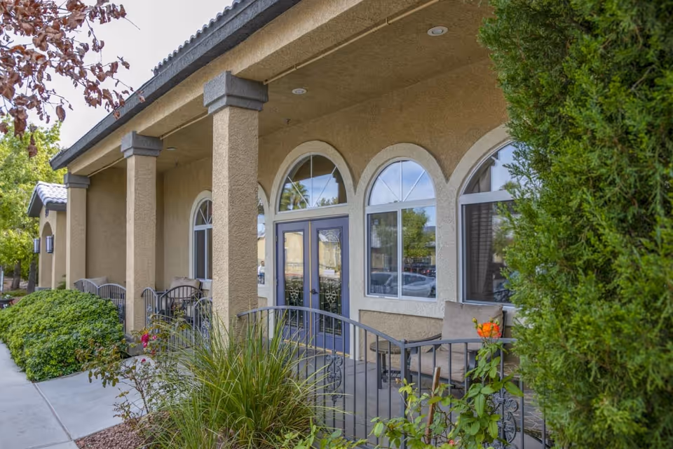 Exterior view of Villa Court Assisted Living showing a beige stucco building with arched windows and a double door entrance. There is a covered porch area with columns, outdoor seating, and surrounding greenery including bushes and trees.