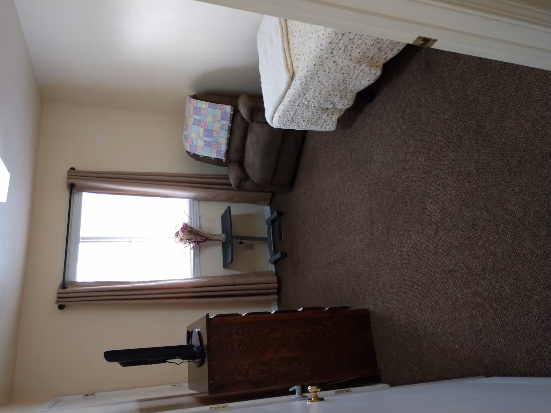 A small bedroom with a single bed covered in a floral bedspread, a brown recliner chair with a colorful patchwork quilt draped over the back, a small black side table with a vase of dried flowers in front of a window with beige curtains, and a wooden dresser with a television on top.