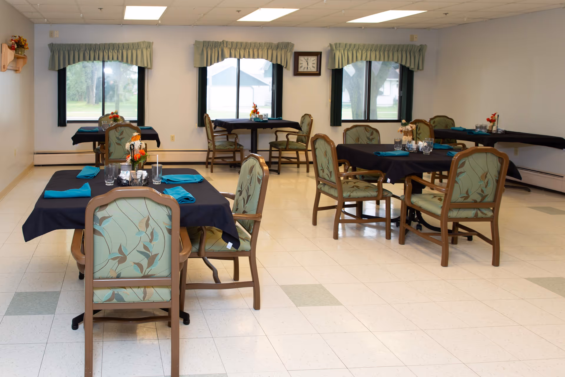 A dining room with several tables covered in black tablecloths and set with turquoise napkins, glasses, and small floral centerpieces. The room has large windows with green valances, light-colored walls, and a tiled floor. Chairs with green patterned upholstery surround the tables.