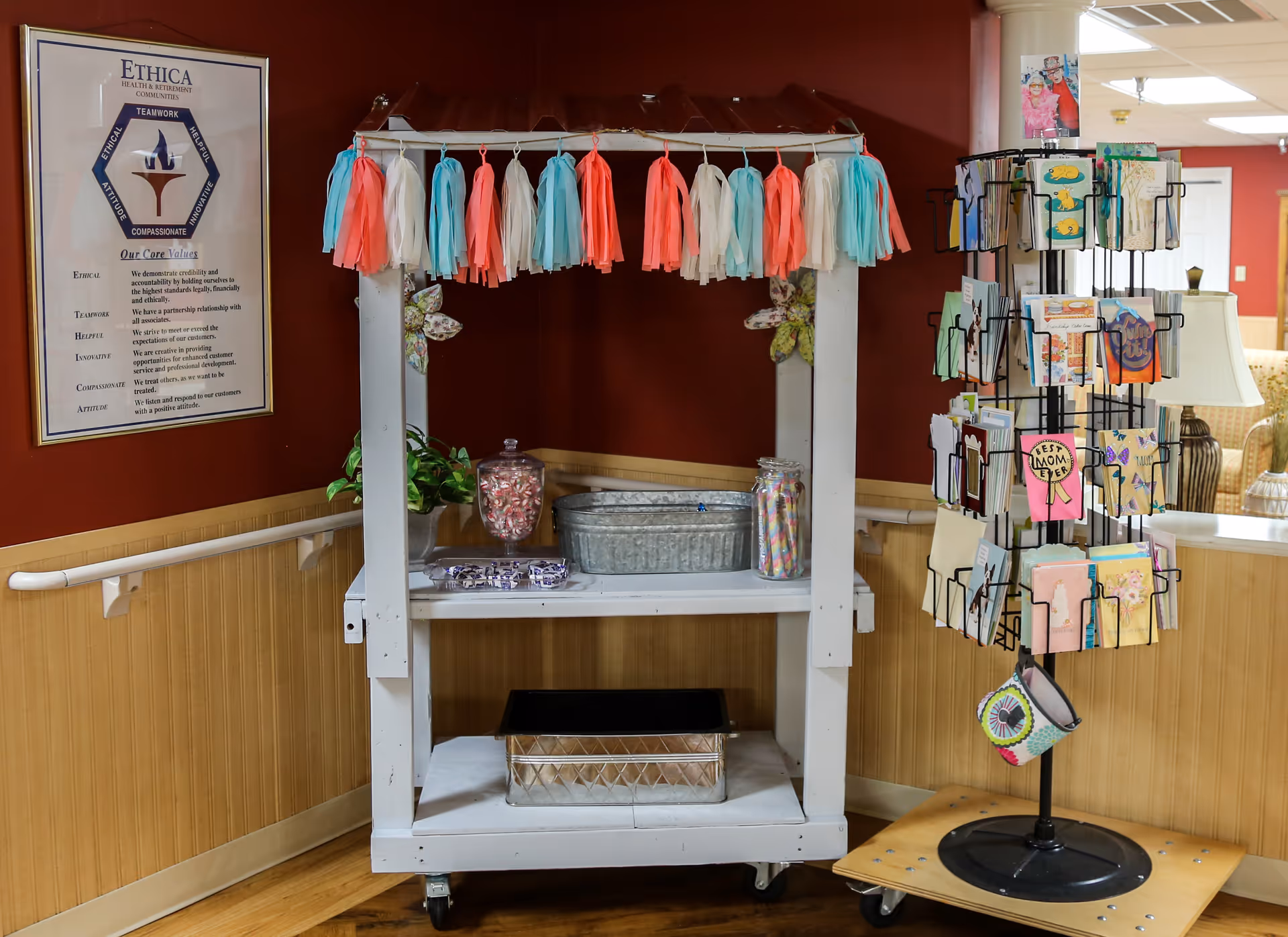 Decorated white display cart with jars of candy and a rotating rack of greeting cards against a red wall in a senior facility common area.