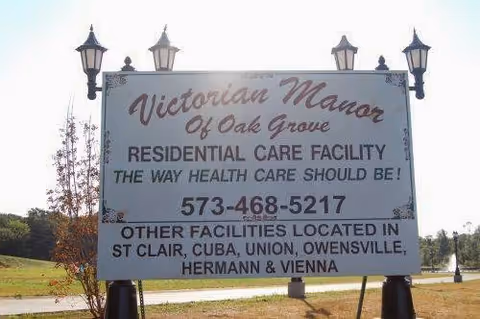 A large outdoor sign for Victorian Manor of Oak Grove Residential Care Facility with contact phone number and a list of other facility locations in St Clair, Cuba, Union, Owensville, Hermann, and Vienna. The sign is mounted on two posts with decorative lanterns on top, set in a grassy area with trees and a road in the background.